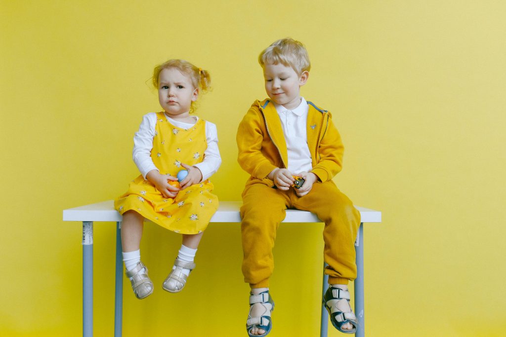 Adorable siblings sitting on a table against a bright yellow background, wearing matching outfits.