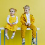 Adorable siblings sitting on a table against a bright yellow background, wearing matching outfits.