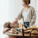 A mother gently wakes her sleepy daughter at a breakfast table filled with healthy foods.